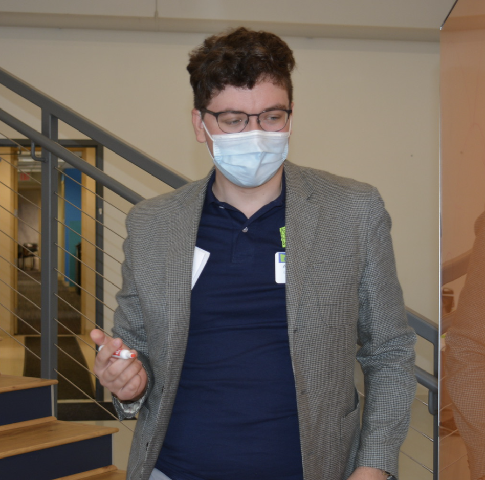 Axel presenting at a student leader training. He is standing up and holding a marker in his hand. He is wearing a surgical mask, glasses, and a medium grey blazer over a blue LeadMN polo
