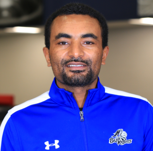 Dame Gamada, is a black man with a short beard and short, curly black hair. He is wearing a royal blue Under Armor zip up and is smiling at the camera. This photo is courtesy of DCTC