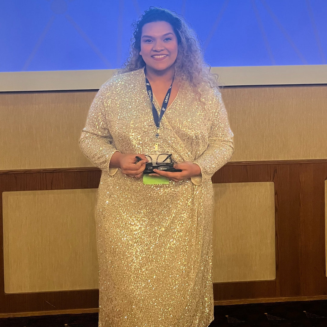 Amona Rite Donald, who is a brown woman with curly blonde hair, smiles at the camera, holding an award at the LeadMN Gala. She is wearing a long sleeve, floor length sparkly cream dress.
