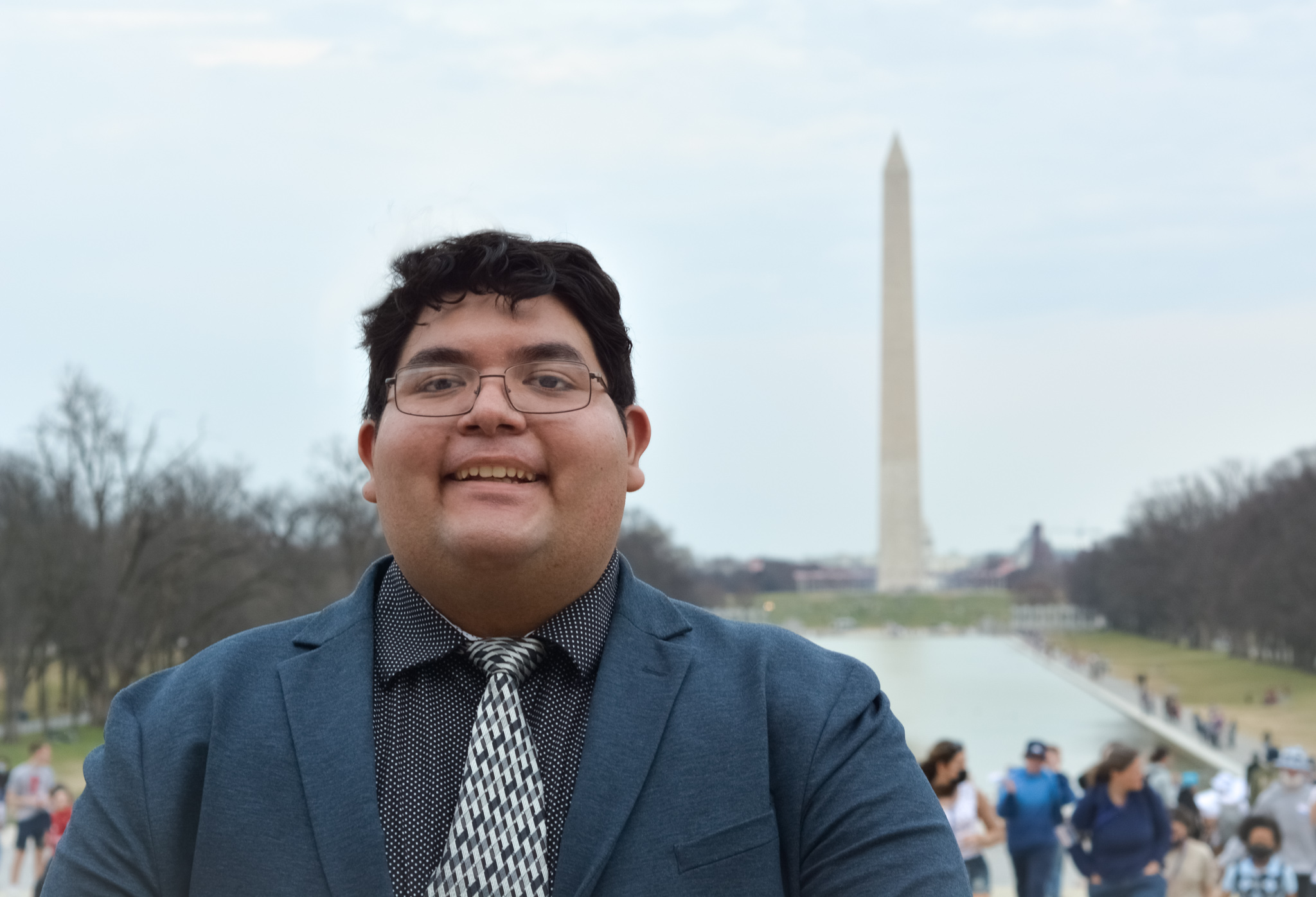 David Mesta, a tall light-skinned brown man with short curly hair and glasses, is smiling in front of the Washington Monument. He is wearing a medium grey suit and a white and grey patterned tie.