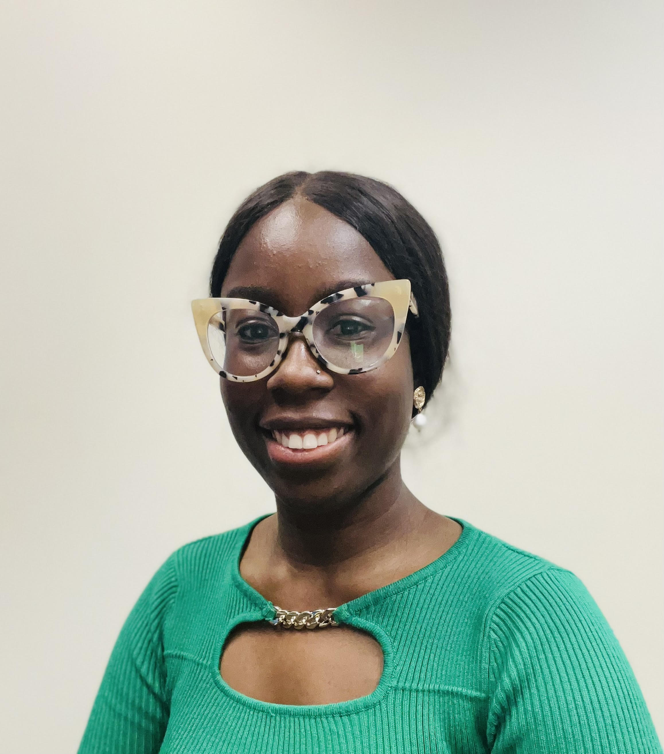Hawa smiles at the camera wearing a grass-green long sleeve top. She is wearing tan and black spotted cat-eye-shape glasses.