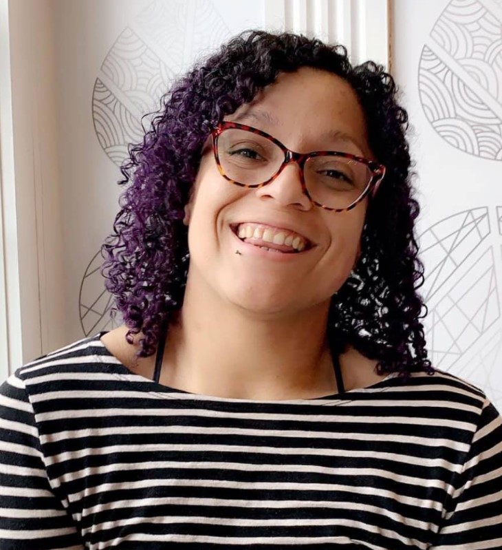 Nezzie Garcia, who is a light-skinned brown woman with curly hair that is black at the top and transitions to purple at the ends, smiles at the camera, wearing a black and white striped shirt. 