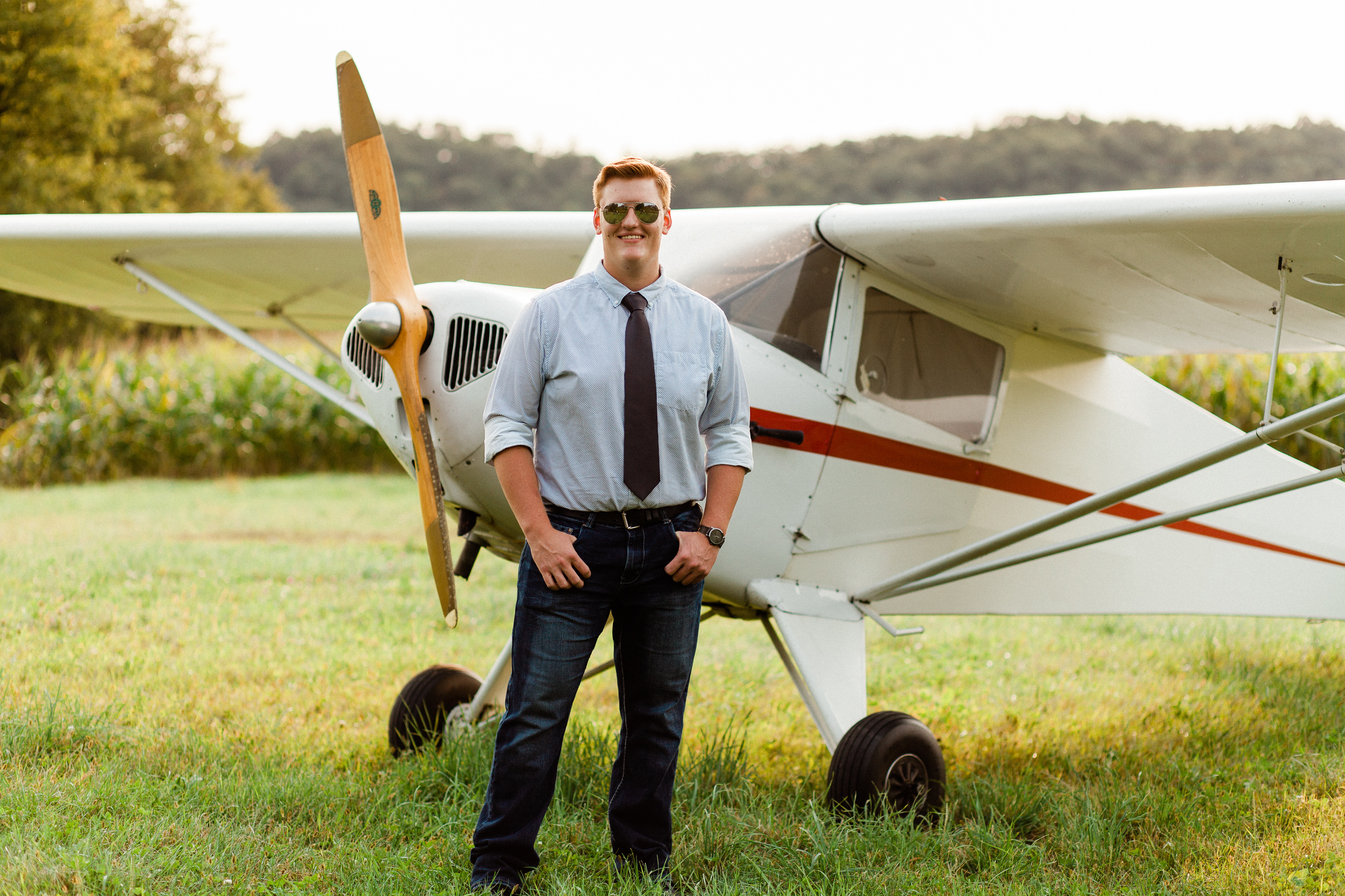 Philip Krzyszton, a white man with red hair, standing next to vintage plane in a green field with green trees. He is wearing aviator sunglasses, dark blue jeans help up with a belt, a light blue button up shirt, and a brown tie.