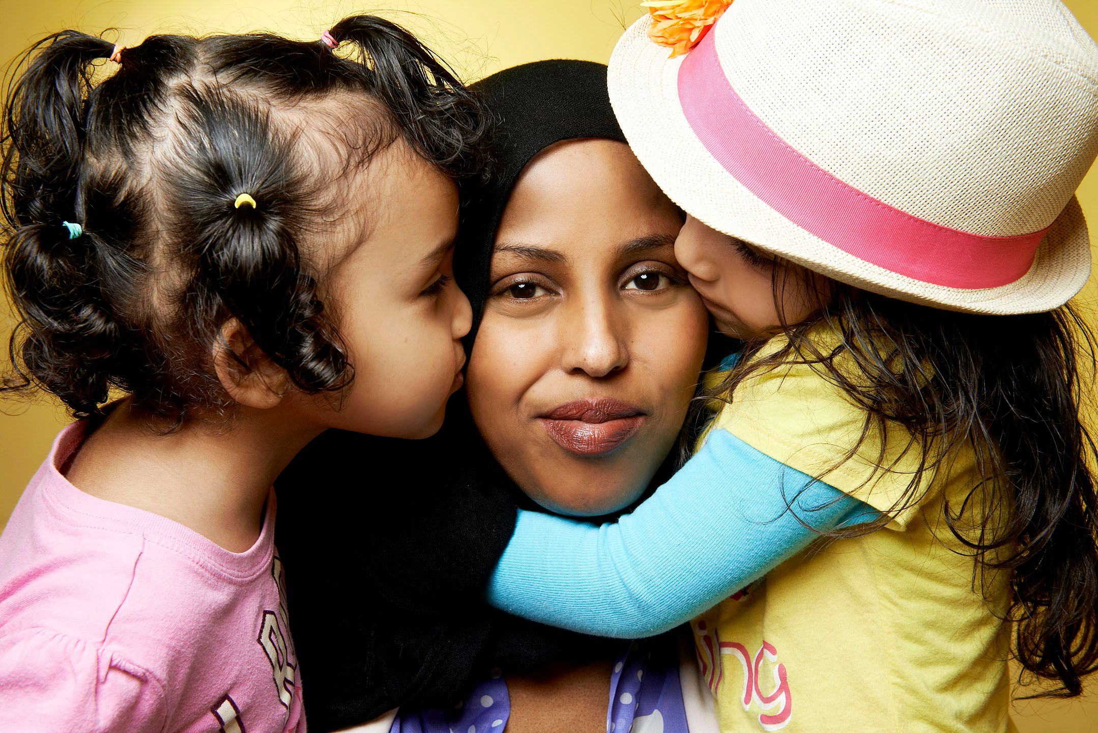 Ladan Abdi, a brown woman with long, straight black hair, smirks at the camera, as she is hugged around the neck by her two young daughters.