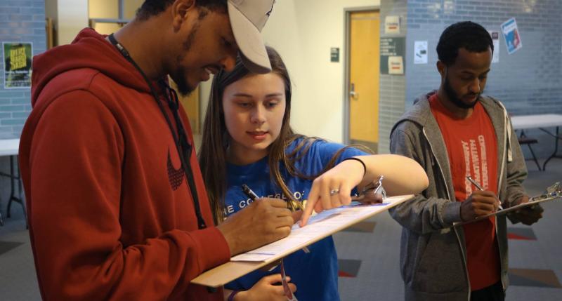 Student with clipboard filling out their registration while another student helps them look over form
