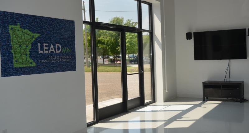 Closed glass doors to the back entrance into the Center for Learning. There is a 3x5 foot poster of the green LeadMN logo on a blue background placed on the wall next to the doors and a TV of a similar size mounted on the wall perpendicular to the doors.