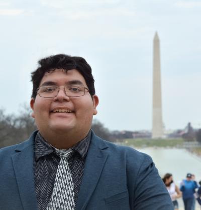 David Mesta, a tall light-skinned brown man with short curly hair and glasses, is smiling in front of the Washington Monument. He is wearing a medium grey suit and a white and grey patterned tie.