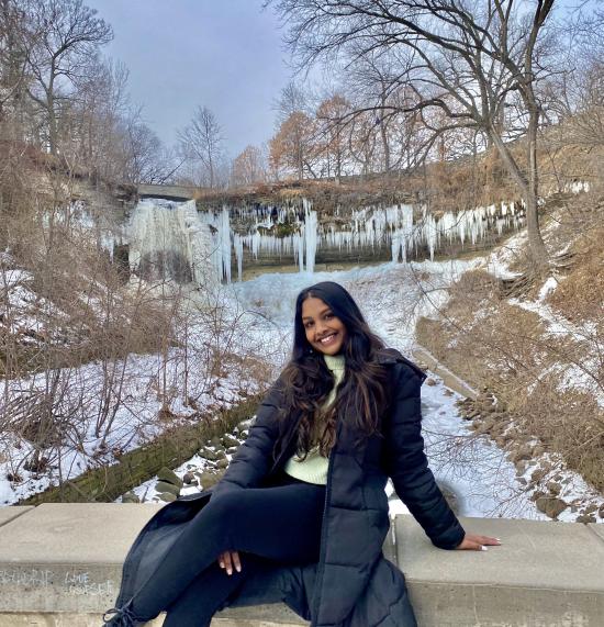 Kimmy Kistan, a brown woman with long, wavy hair that is black with a balayage fade into medium-brown. She wearing an unzipped calf-length puffy black jacket on top of a green sweater and black leggings. She is smiling at the camera, posing in front of a snowy nature background.