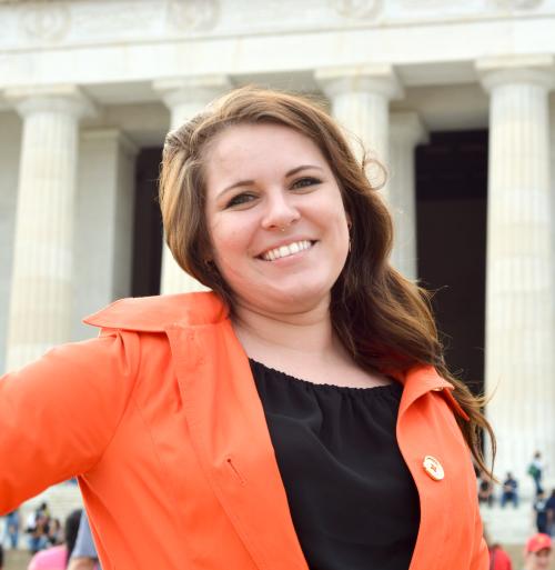 Ali Tomashek, a white woman with medium brown hair, smiles in front of Lincoln Memorial in bright orange coat.
