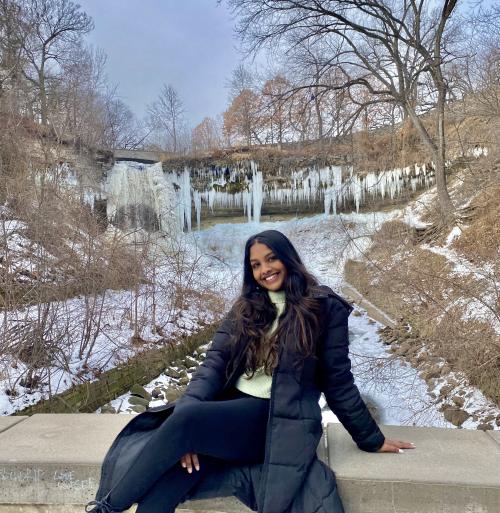 Kimmy Kistan, a brown woman with long, wavy hair that is black with a balayage fade into medium-brown. She wearing an unzipped calf-length puffy black jacket on top of a green sweater and black leggings. She is smiling at the camera, posing in front of a snowy nature background.