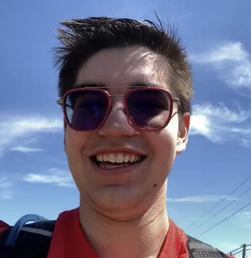Caledon Johnston, a white man with short brown hair, smiles for a selfie. Ice is wearing red-framed aviator sunglasses and it is a sunny day.