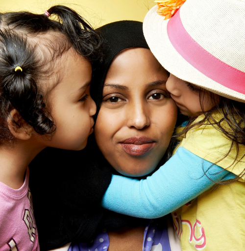 Ladan Abdi, a brown woman with long, straight black hair, smirks at the camera, as she is hugged around the neck by her two young daughters.